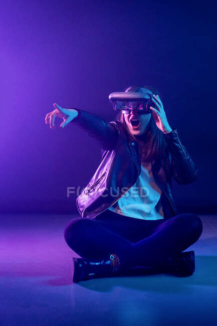 Неузнаваемая женщина с вытянутой рукой в VR гарнитуре, исследуя виртуальную реальность под синим неоновым светом возле стены с освещением проектора — стоковое фото