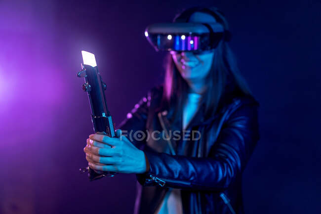 Anonymous smiling female wearing modern VR headset standing in dark room with light saber with white neon light in hands while exploring virtual reality — Stock Photo
