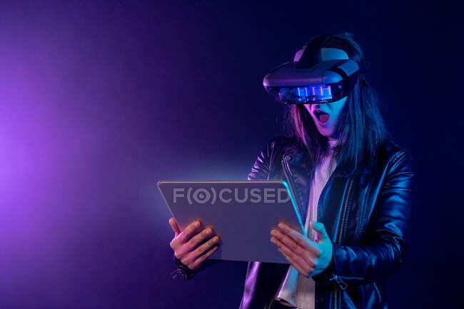 Donna anonima concentrata con auricolare VR moderno in piedi vicino alla parete in camera buia con tablet contemporaneo in mano sotto illuminazione al neon — Foto stock