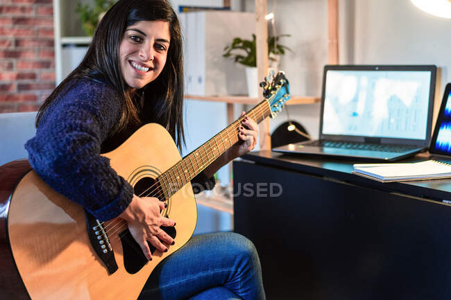 Vista laterale della chitarra acustica femminile mentre componeva musica vicino al tavolo con laptop in camera con muro di mattoni durante il lavoro a distanza — Foto stock