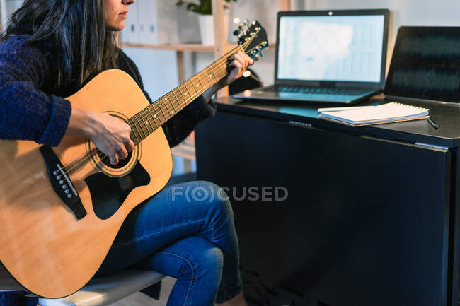 Vista lateral da fêmea tocando guitarra acústica ao compor música perto da mesa com laptop no quarto com parede de tijolo durante o trabalho remoto — Fotografia de Stock