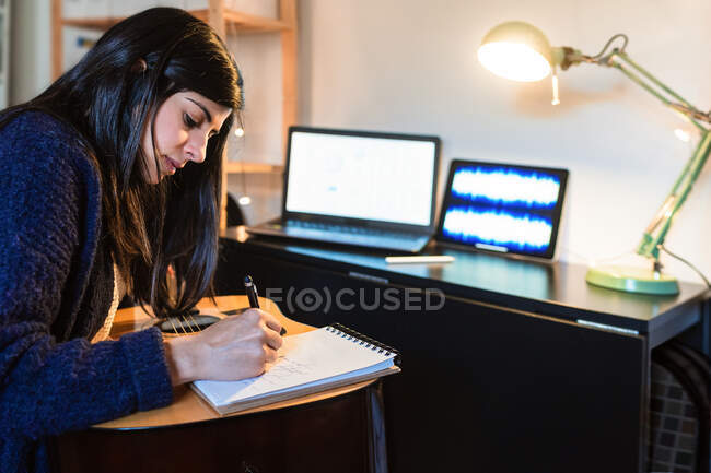 Вид збоку концентрованої жінки-композитора, що робить нотатки в блокноті, сидячи за столом з ноутбуком під час віддаленої роботи з дому — стокове фото