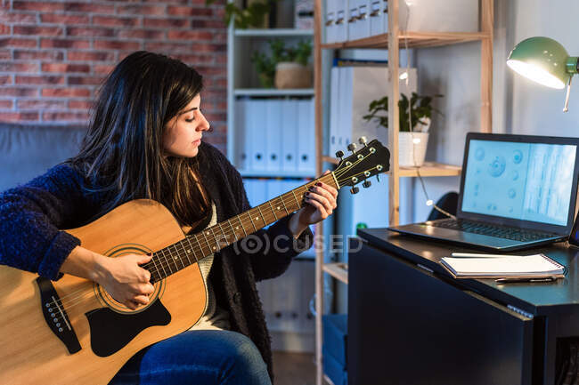 Side view of female playing acoustic guitar while composing music near table with laptop in room with brick wall during remote work — Stock Photo