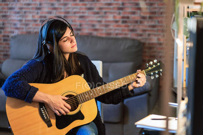 Femmina suonare la chitarra acustica durante la composizione di musica vicino al tavolo con computer portatile in camera con muro di mattoni durante il lavoro a distanza — Foto stock