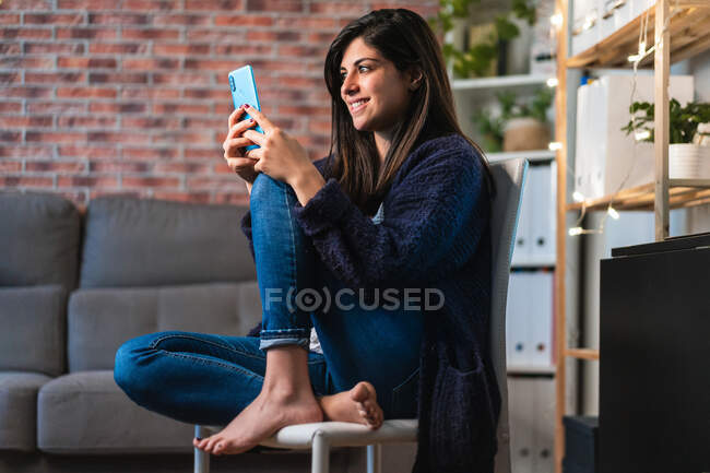 Positive Freiberuflerin surft mit dem Handy und sitzt neben Tisch und Regalen mit Dekorationen, während sie von zu Hause aus arbeitet — Stockfoto
