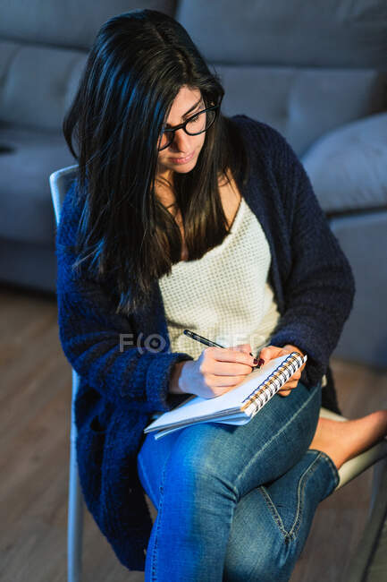 Зверху концентрований жіночий композитор робить нотатки в блокноті, сидячи за столом з ноутбуком під час віддаленої роботи з дому — стокове фото