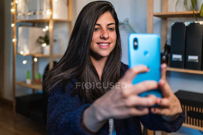 Positivo freelance femminile che si fa selfie e si siede vicino a tavolo e scaffali con decorazioni mentre lavora a distanza da casa — Foto stock