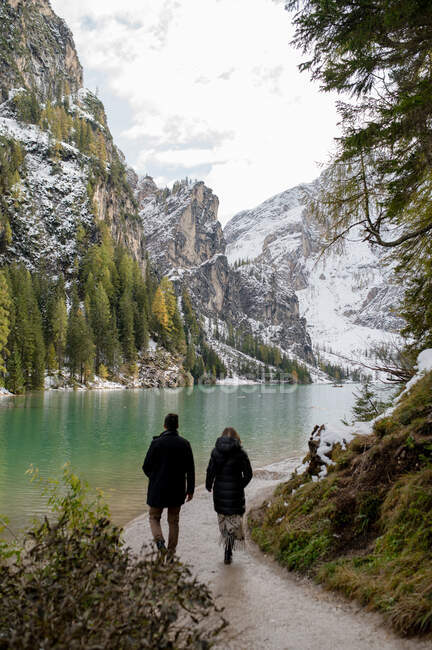 Vue arrière d'un couple méconnaissable en vêtements de dessus marchant le long du lac Lago di Braies dans les hautes terres d'Italie — Photo de stock
