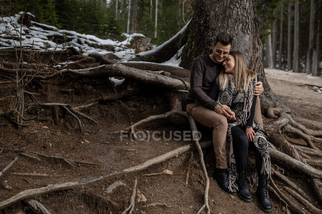 Full body of positive gentle couple sitting near tree and hugging each other during romantic day in woods — Stock Photo