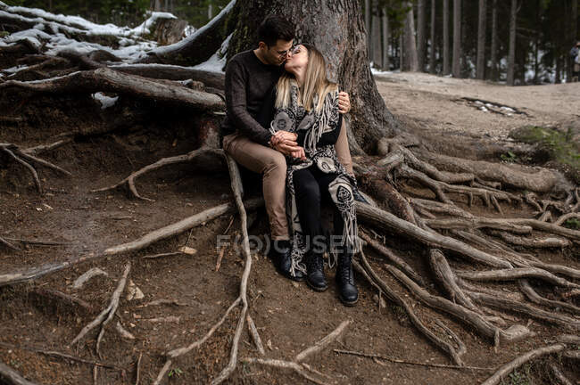 Full body of positive gentle couple sitting near tree and hugging and kissing each other during romantic day in woods — Stock Photo