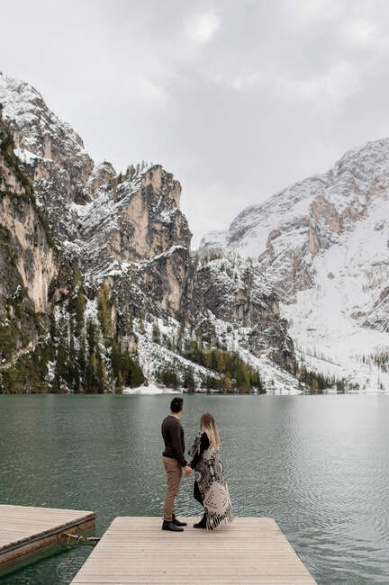 Corps complet d'un couple aimant et doux s'embrassant sur une jetée en bois contre le lac Lago di Braies entouré de montagnes enneigées — Photo de stock