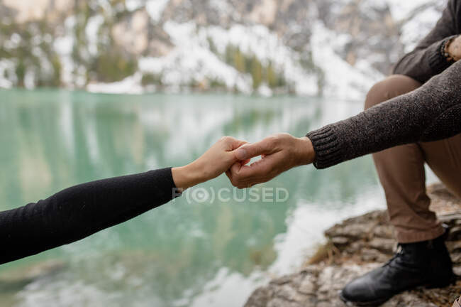 Crop anonymous traveler holding hand with girlfriend while supporting for climbing on rocky shore of Lago di Braies lake in Italy — Stock Photo