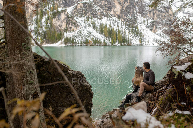 Full body of loving gentle couple hugging each other while sitting on woods against Lago di Braies lake surrounded by snowy mountains — Stock Photo