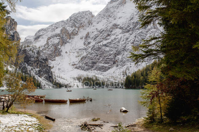 Paisaje de barcos de madera amarrados en un tranquilo lago ondulado rodeado de montañas nevadas y árboles de coníferas - foto de stock