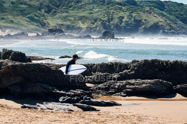 Side view of male tourist with surfboard walking along boulders on sandy coast of waving foaming sea — Stock Photo