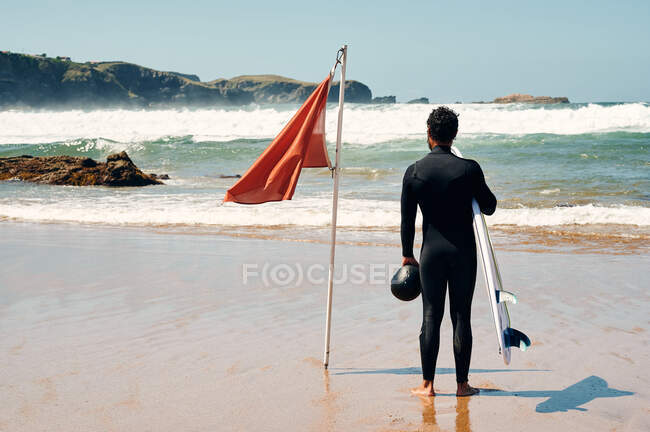Vista posteriore piena lunghezza dell'uomo anonimo che indossa tuta da surf in piedi vicino alla bandiera sventolante sulla costa lavata da onde schiumose — Foto stock