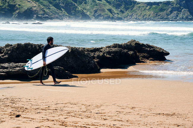 Adult man in wetsuit with surfboard smiling happily standing on seashore against hills — Stock Photo