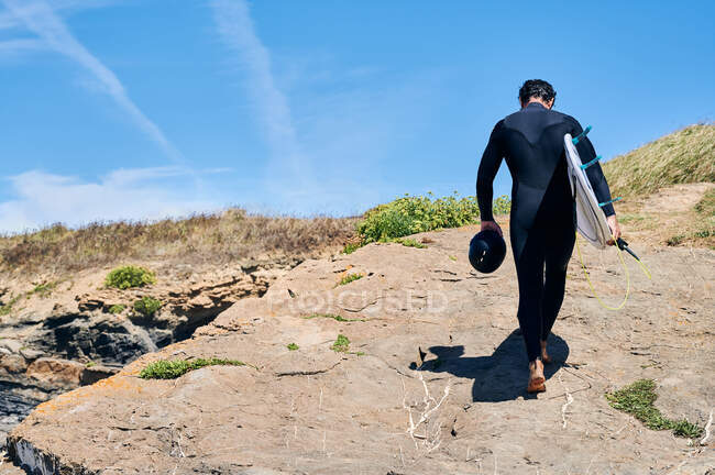Back view full length of anonymous man wearing surfing suit standing near waving flag on coast washed by foamy waves — Stock Photo