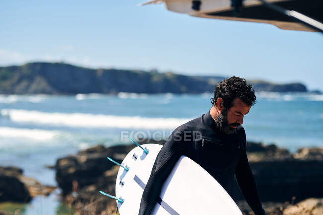 Hombre adulto en traje de neopreno con tabla de surf sonriendo felizmente de pie en la orilla del mar contra las colinas - foto de stock