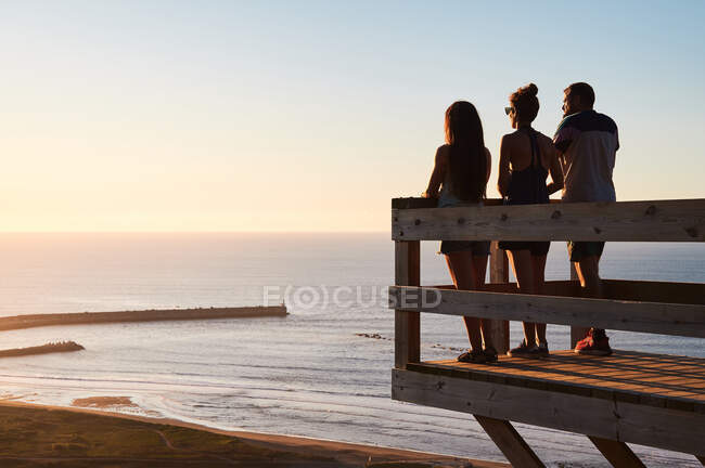 Full body back view of tourists standing on wooden platform and admiring calm sea during sundown — Stock Photo