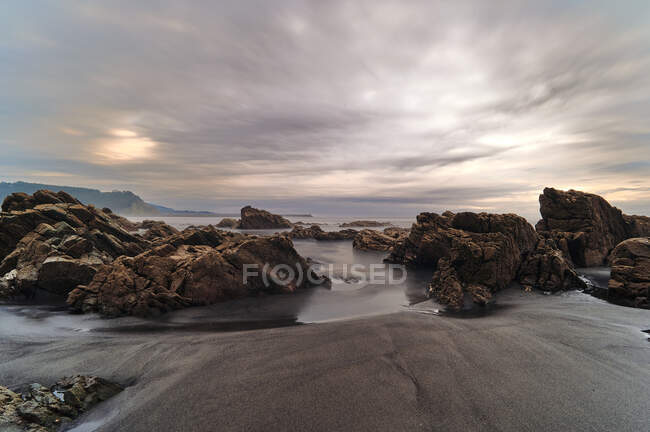 Pintoresca vista de rocas cubiertas de musgo en la playa de arena de mar bajo el cielo nublado del atardecer - foto de stock