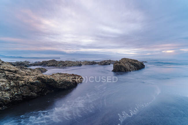 Pintoresca vista de rocas cubiertas de musgo en la playa de arena de mar bajo el cielo nublado del atardecer - foto de stock