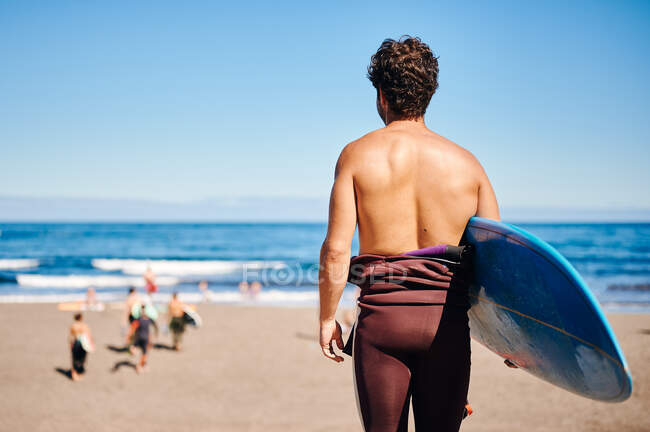 Vista posterior de un atleta masculino irreconocible con tabla de surf admirando el mar ondeante en un día soleado sin nubes - foto de stock