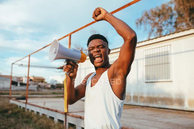 Joven afroamericano enojado en camiseta con altavoz gritando con el brazo levantado mientras mira a la cámara - foto de stock
