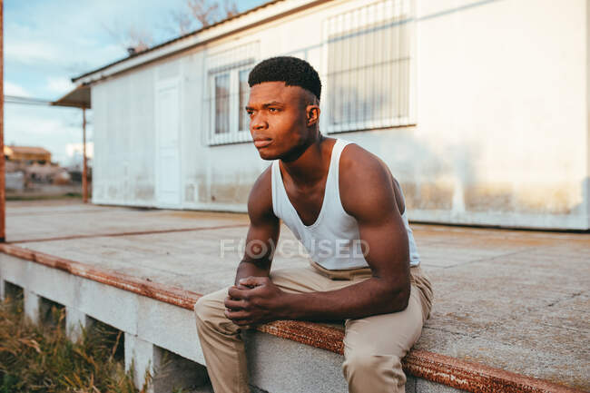 Joven masculino afroamericano en camiseta con las manos cerradas mirando hacia otro lado contra la casa - foto de stock