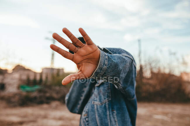 Anonymous ethnic male in denim jacket hiding face with reached arm under cloudy sky in daylight — Stock Photo