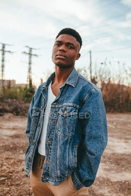 Giovane maschio afroamericano serio con taglio di capelli moderno in giacca di jeans guardando la fotocamera sotto il cielo nuvoloso in autunno — Foto stock