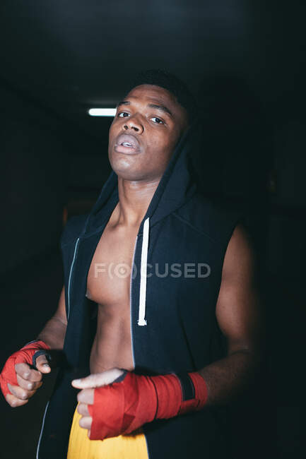 Молодой сильный афроамериканский спортсмен в боксёрских обертках для рук работает и смотрит на камеру в здании — стоковое фото