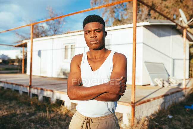Joven masculino afroamericano en camiseta de pie cerca de la plataforma y mirando a la cámara contra la casa - foto de stock