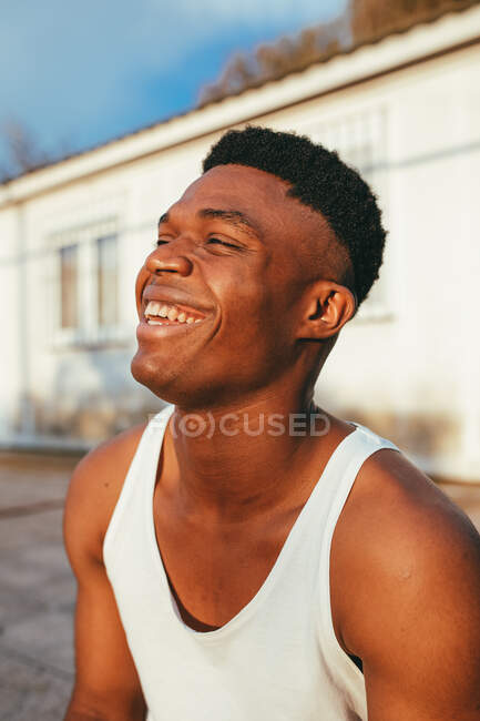 Homem afro-americano feliz em camiseta com corte de cabelo moderno olhando para a frente contra a construção à luz do sol — Fotografia de Stock