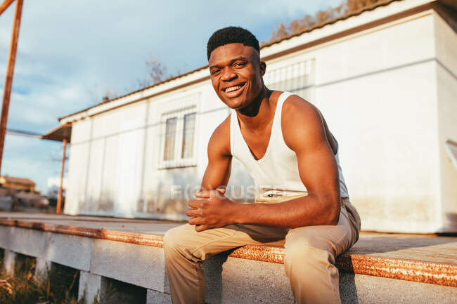 Homem afro-americano feliz em camiseta com corte de cabelo moderno olhando para a câmera contra a construção à luz do sol — Fotografia de Stock