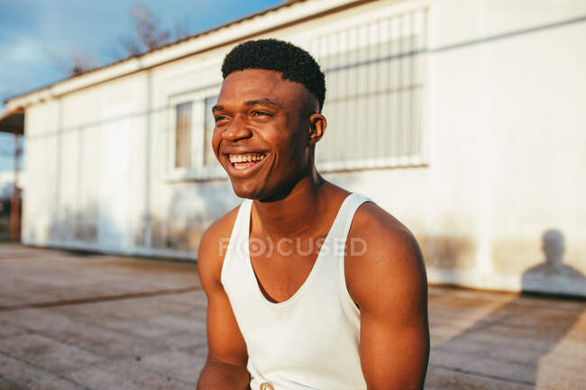 Hombre afroamericano feliz en camiseta con corte de pelo moderno mirando hacia adelante contra la construcción en la luz del sol - foto de stock