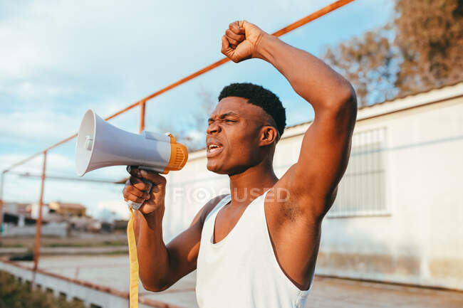 Jovem afro-americano irritado macho em camiseta com alto-falante gritando com braço levantado enquanto olha para a câmera — Fotografia de Stock