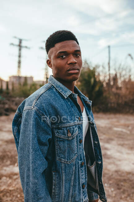 Молодий афроамериканець з сучасною зачіскою в джинсовій куртці дивиться на камеру під хмарним небом. — стокове фото