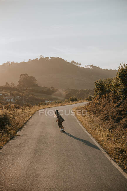 Back view faceless sporty female in trendy wear riding cruiser board along empty asphalt road in summer countryside on sunny day — Stock Photo