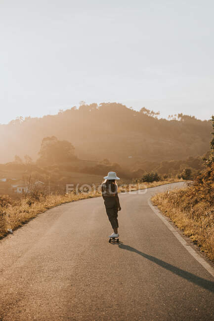 Вид сзади безликая спортивная женщина в модной одежде на крейсерской доске вдоль пустой асфальтовой дороги в летней сельской местности в солнечный день — стоковое фото