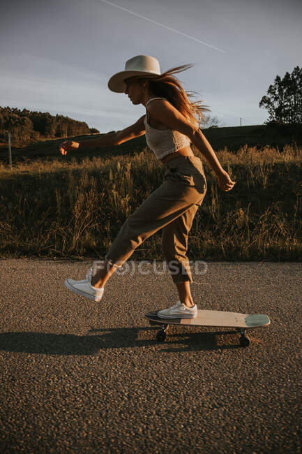 Side view sporty female in trendy wear riding cruiser board along empty asphalt road in summer countryside on sunny day — Stock Photo