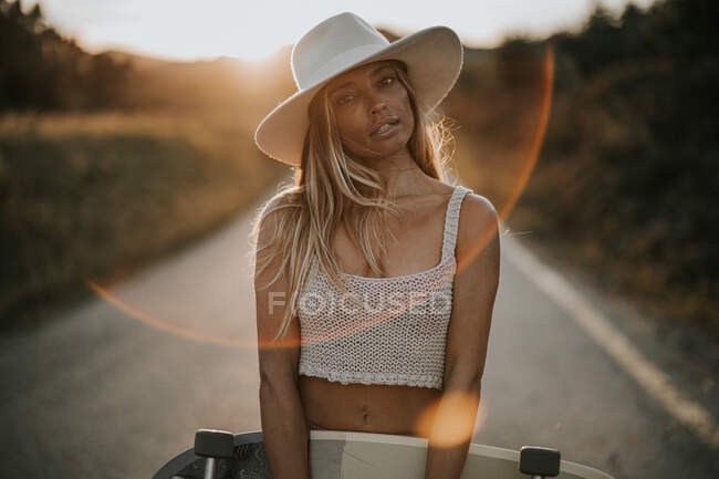 Mulher nova no desgaste casual e chapéu de verão que prende o skate do cruzador e que olha para a câmera quando estando na estrada vazia do asfalto na área rural no por do sol — Fotografia de Stock