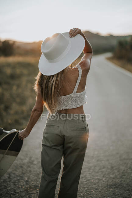 Вид ззаду молода жінка в повсякденному вбранні і літньому капелюсі тримає крейсер скейтборд і дивиться в сторону, стоячи на порожній асфальтовій дорозі в сільській місцевості на заході сонця — стокове фото