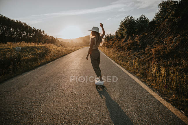 Вид сзади Спортивная женщина в модной одежде верхом на крейсерской доске вдоль пустой асфальтовой дороги в летней сельской местности в солнечный день — стоковое фото