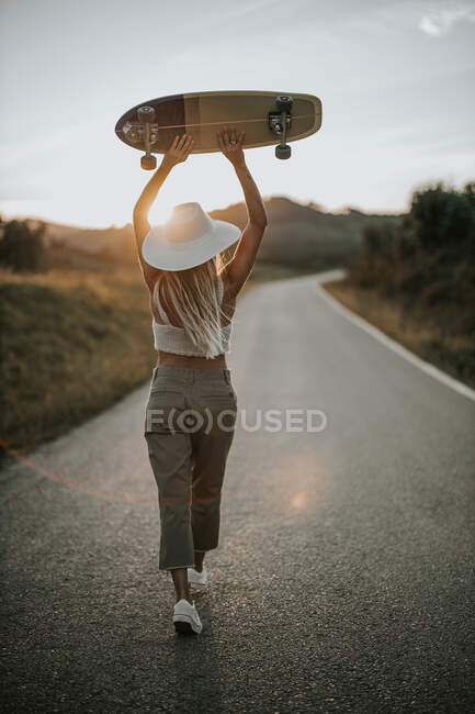 Смотреть назад молодая женщина в повседневной одежде и летняя шляпа держа крейсер скейтборд и глядя в сторону, стоя на пустой асфальтовой дороге в сельской местности на закате — стоковое фото