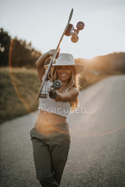 Conteúdo jovem fêmea em desgaste casual e chapéu de verão segurando skate cruzador e olhando para a câmera enquanto estava em pé na estrada de asfalto vazio na área rural ao pôr do sol — Fotografia de Stock