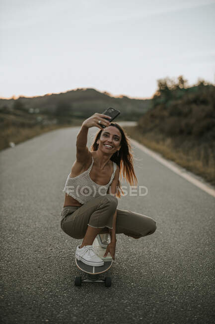 Pieno corpo attraente spensierato pattinatore femminile seduto su fianchi sullo skateboard e prendendo selfie mentre pattina sulla strada rurale vuota al crepuscolo — Foto stock