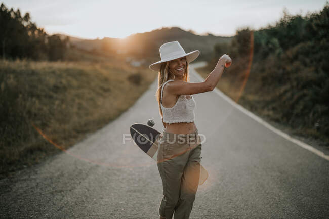 Contenida joven hembra en ropa casual y sombrero de verano sosteniendo el monopatín cruiser y mirando a la cámara mientras está parada en un camino de asfalto vacío en el área rural al atardecer - foto de stock