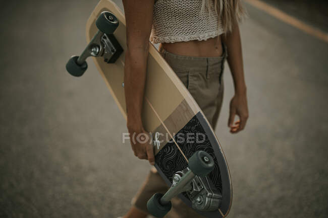 Crop young female in casual wear holding cruiser skateboard while standing on empty asphalt road in rural area at sunset — Stock Photo