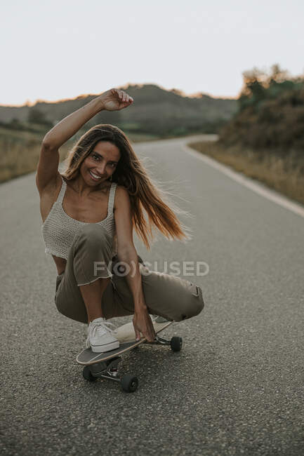 Полнотелая привлекательная беззаботная фигуристка сидит на ягодицах на скейтборде и смотрит в камеру, катаясь на коньках по пустой сельской дороге в сумерках — стоковое фото